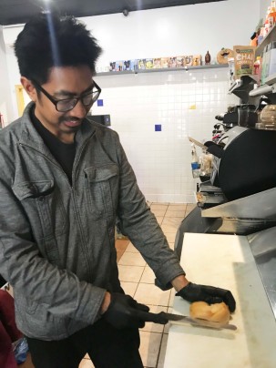 Ryan Natividad wears a gray jacket and gloves. He slices a bagel in the kitchen of his shop, The Bagel Exchange. 