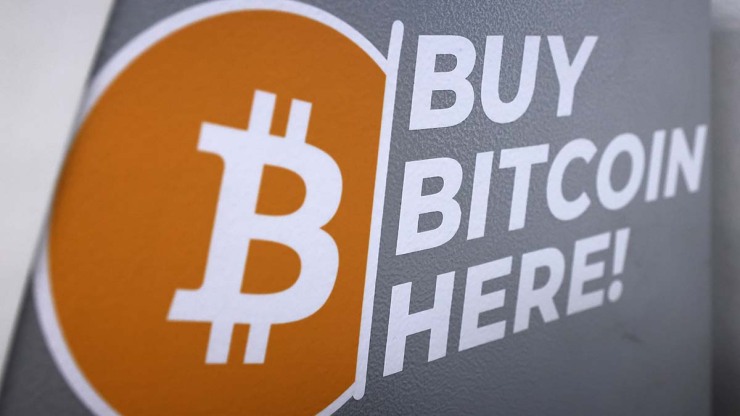 The side of a Bitcoin ATM displays an orange and white bitcoin logo (a B with two vertical lines through it like a dollar sign) in an orange circle, with white text reading 
